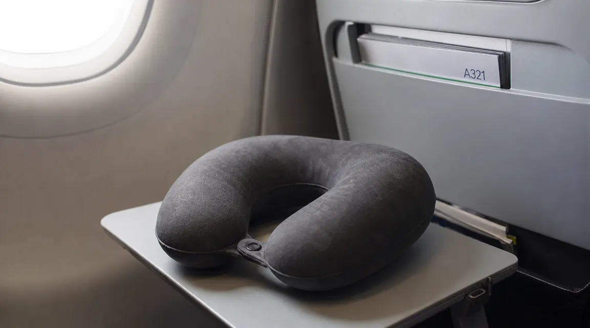 Travel neck pillow resting on an airplane tray table next to a window, soft daylight streaming through the cabin